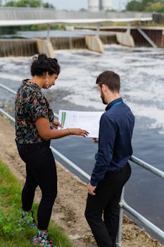 Male and female engineers reviewing plans outdoors by a river dam.